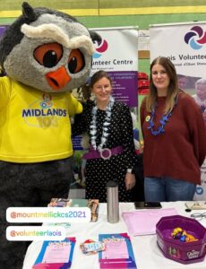 Laois Volunteer Centre team (Emer & Catriona, right to left) standing at their table at the Ogra Live Well Expo next to the mascot for Midlands 103